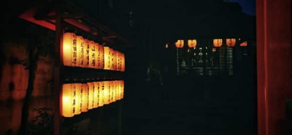 Des lampions allumés avec des kanjis inscrits dessus. On distingue les contours d'un temple dans le noir.